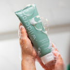 Soapy, wet hands hold a green tube of Wildcraft shampoo covered in white suds and iridescent bubbles.