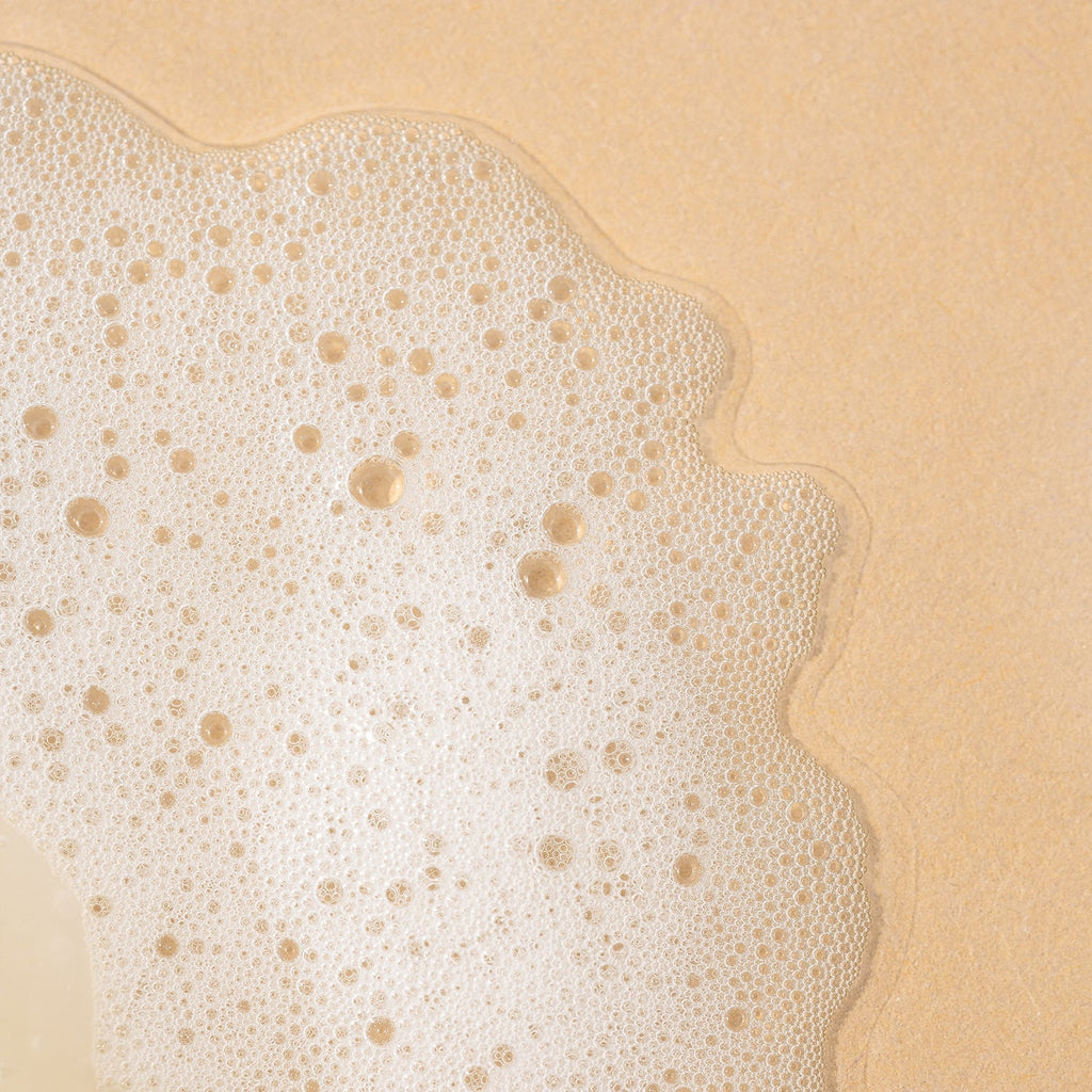 A close-up texture shot of fine white soapy lather and bubbles spread over a textured beige surface.