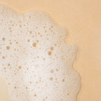 A close-up texture shot of fine white soapy lather and bubbles spread over a textured beige surface.