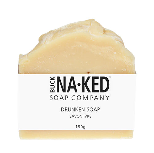 An unevenly cut beige bar soap wrapped in a white Buck Naked Soap Company paper label against a white background.