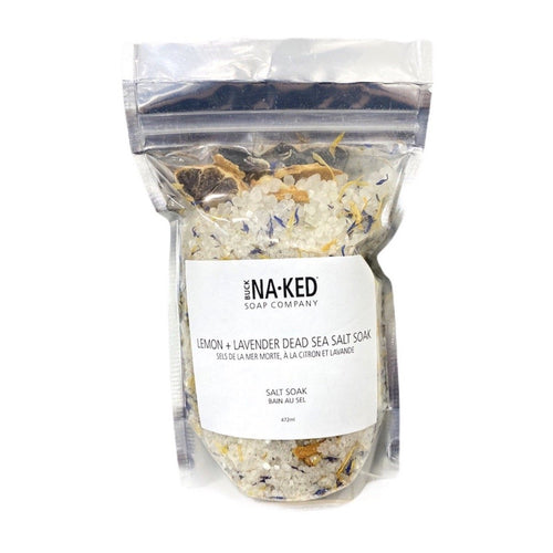 A clear pouch of Buck Naked Lemon + Lavender Dead Sea Salt Soak, showing coarse white crystals and dried flower petals.